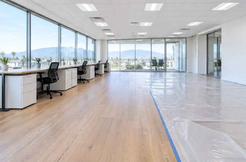 Illustration: a bright open office in Ontario, CA with a wide oak plank floor half covered by plastic sheeting for a phased refinish and desks pushed against the glass wall