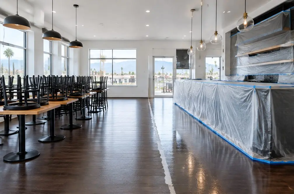 Illustration: a dining room in Ontario, CA with a dark-stained oak floor, empty tables stacked to one side, and plastic over the bar while the floor is being recoated before opening